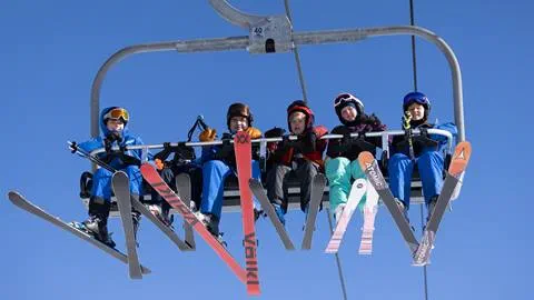 Skiers wearing helmets and goggles seated on a chairlift with their skis dangling below.