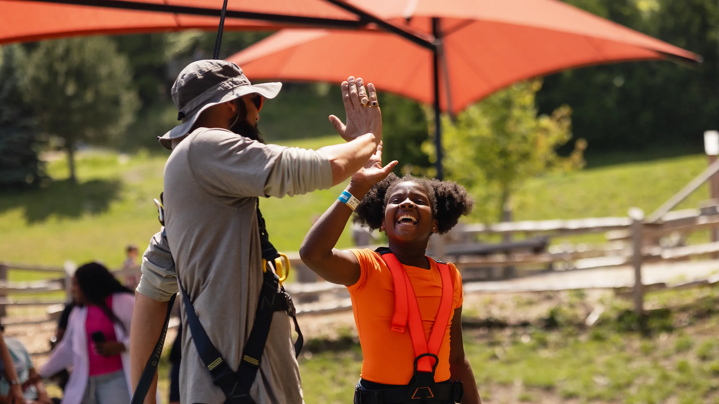 A child wearing a harness gives a high five to an instructor outdoors.