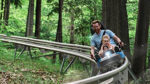 A man and a small child riding on a single-car roller coaster through a forested area, both wearing safety harnesses.