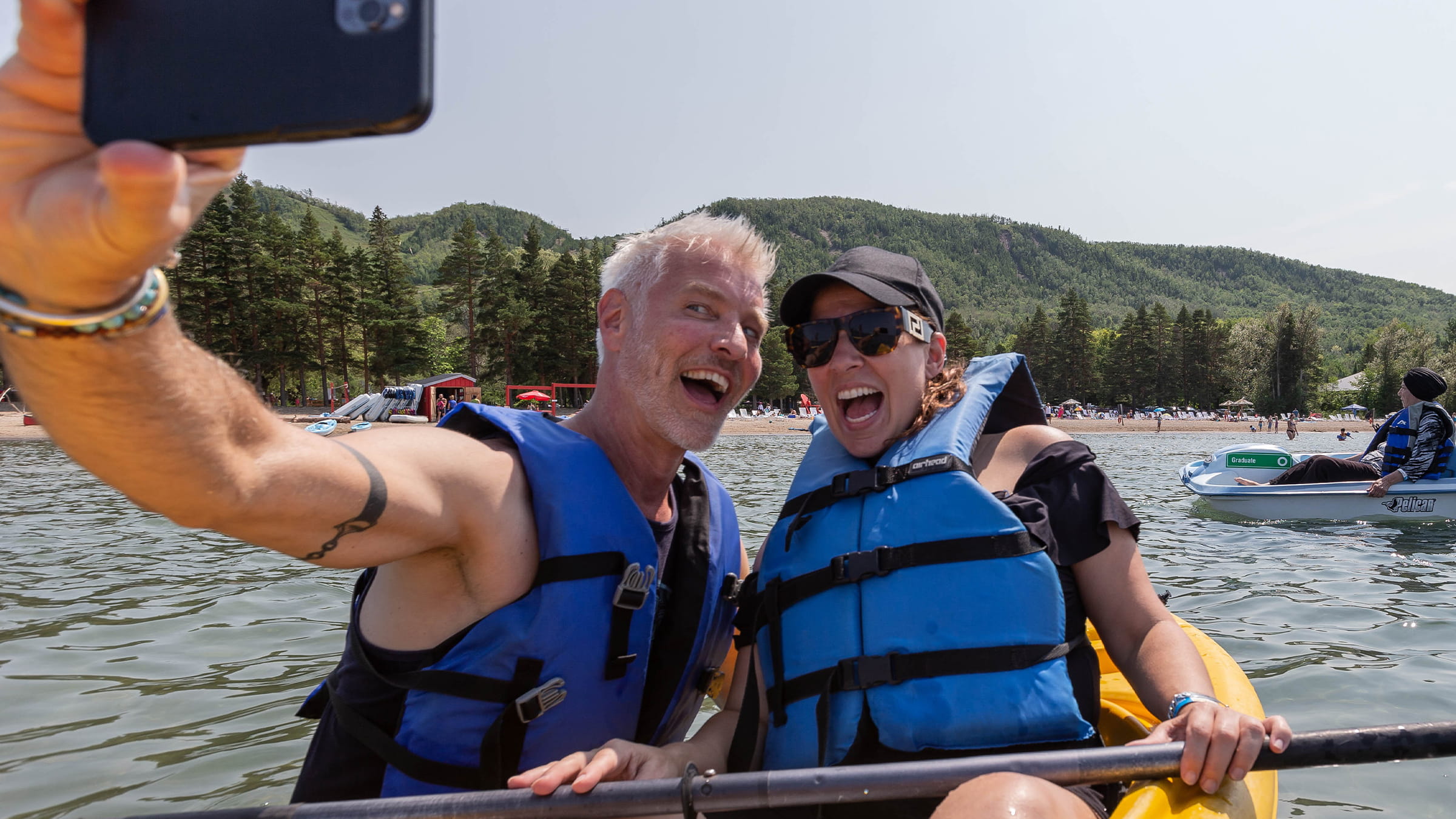 Two people in blue life jackets sitting in a kayak, taking a selfie with a smartphone, smiling widely, with a scenic background of green hills and trees.