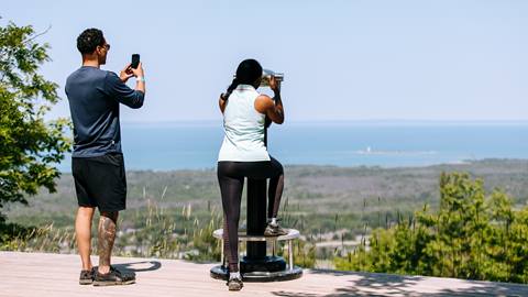 A man and a woman on an elevated lookout; he is taking a photo with his phone, she is using a viewing binocular, with a coastal landscape in the background.