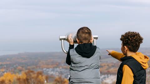 Two children are outdoors, looking through a telescope. One child is pointing toward the distance while the other peers through the eyepiece. Trees with autumn foliage are visible in the background.