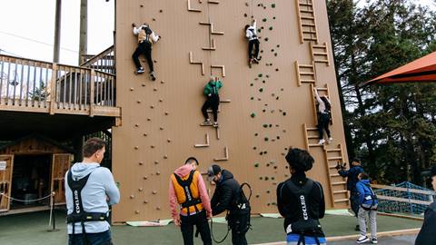 Several people are climbing a large outdoor rock climbing wall while others watch and prepare with harnesses at the base. The wall features different climbing routes and is surrounded by trees.