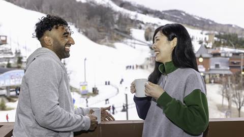 A man and woman talking on a balcony overlooking a ski resort.