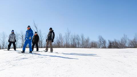 Four people wearing winter gear stand on skis and snowboards on a snowy slope under a clear blue sky with leafless trees in the background.