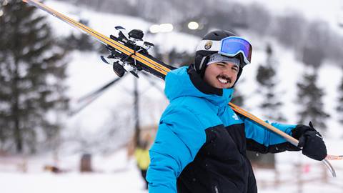 A person in a blue and black jacket and ski goggles smiles while carrying skis on their shoulder on a snowy slope. Trees and a snowy landscape are visible in the background.