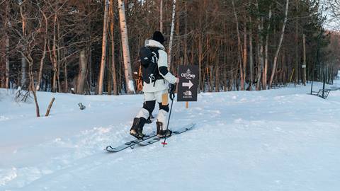 Two people on a snowy trail using skis and carrying backpacks, following a sign pointing to a destination. Trees line the path.