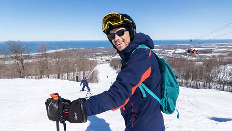 A person in ski gear and a backpack stands on a snowy slope, smiling, with a ski pole. Another skier is seen descending in the background under a clear blue sky.