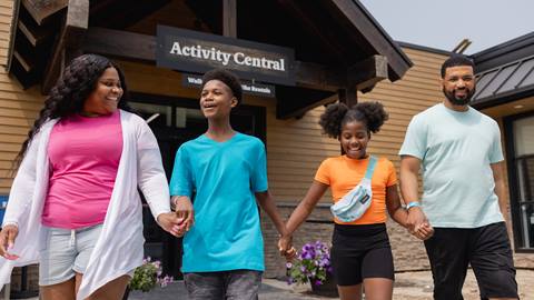 a family of four walking hand in hand near the "activity central" building, smiling and enjoying themselves at blue mountain resort