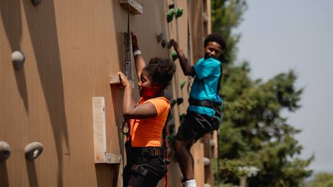 Two children wearing safety harnesses climb a wooden rock wall. The girl in orange is in the foreground, the boy in blue is slightly behind her. Trees and sky are visible in the background.
