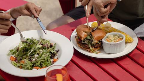 Two people eating outdoors at a red table: one has a salad, the other has a burger, corn on the cob, and coleslaw. A glass with a beverage is in the foreground.