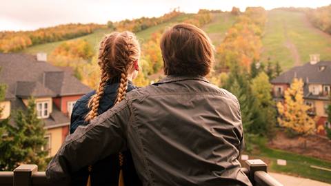 Two people with braided hair and jackets stand on a balcony, looking at a hilly landscape with autumn foliage and houses below.