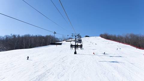 A ski lift carries skiers up a snowy slope with several people skiing down under a clear blue sky. The slope is surrounded by some trees on the sides.