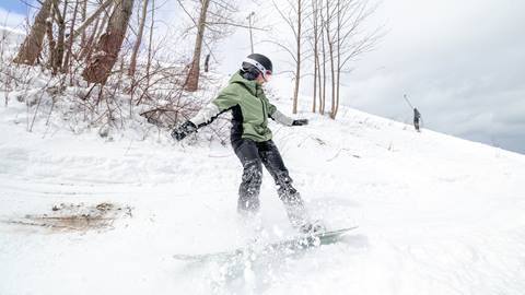 Person snowboarding downhill on a snowy slope, surrounded by bare trees and a cloudy sky.