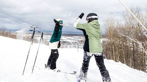 Two snowboarders on a snowy slope high-five each other. Ski poles are planted in the snow nearby. Trees and a distant view of a town are visible in the background.