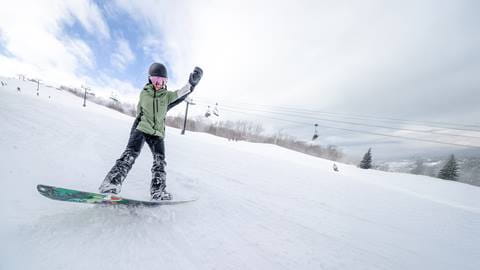 A snowboarder in a green jacket and black pants glides down a snowy slope with one arm raised. Ski lifts and leafless trees are visible in the background under a partly cloudy sky.