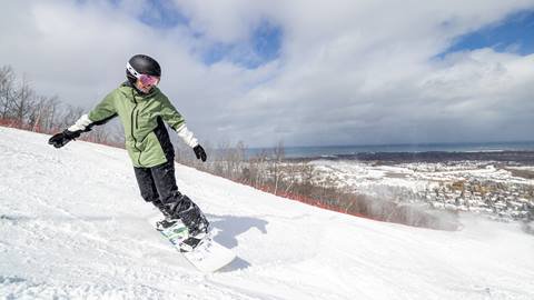 Person wearing a green jacket and black helmet snowboarding down a snowy mountain slope on a clear day with a view of a town and body of water in the background.