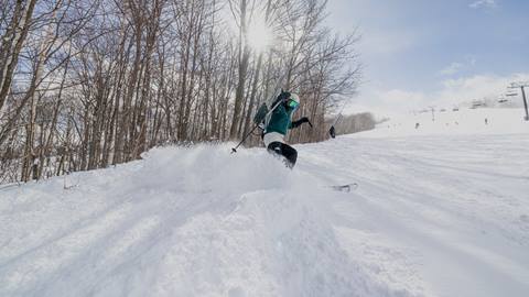 A skier descends a snowy slope under a sunny sky, with trees on the left and a ski lift in the background.