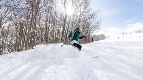 A skier descends a snowy slope under a sunny sky, with trees on the left and a ski lift in the background.