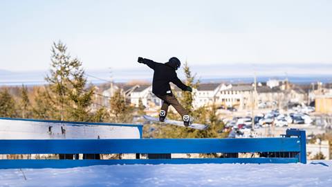 A person wearing a black helmet and jacket snowboards on a rail in a snowy, outdoor setting with buildings and trees in the background.