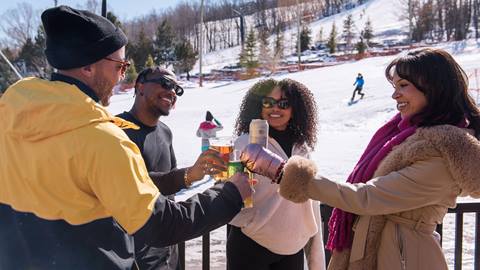 Four people wearing winter clothing toast with drinks outdoors, with a snowy ski slope in the background.