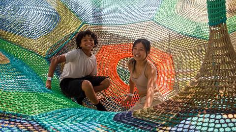 Two children smile while sitting inside a colorful, netted play structure made of interwoven ropes.