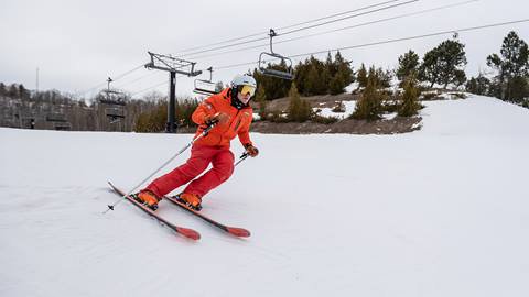 A person in an orange jacket is skiing downhill on a snowy slope with chairlifts in the background.