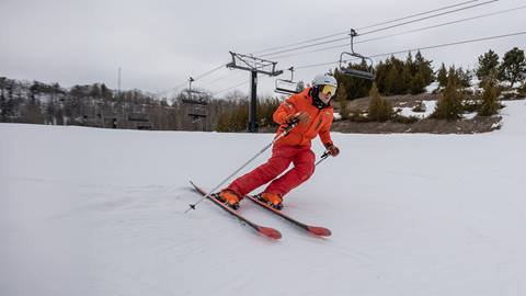 A person in an orange jacket is skiing downhill on a snowy slope with chairlifts in the background.