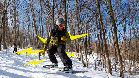 Snowboarder performing a jump on a snowy slope with another individual skiing in the background.