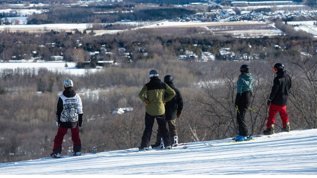 Skiers on a snowy hilltop overlooking a valley.