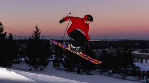 A snowboarder in a red jacket and black pants performs a jump against a sunset sky at a snowy resort.