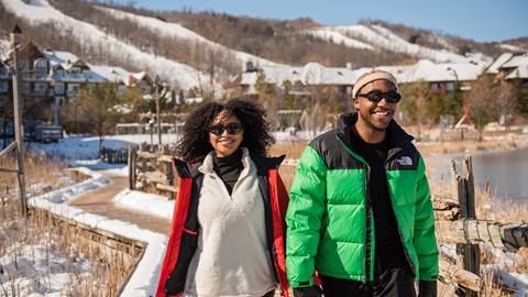 Two people wearing winter jackets walk along a snowy path with mountains and buildings in the background on a sunny day.