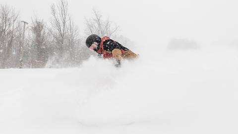 Person snowboarding through deep powder on a snowy day, surrounded by trees in the background.