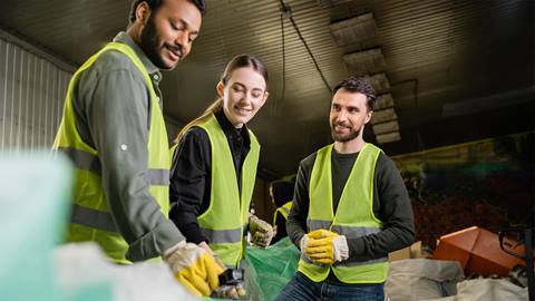 Three workers in reflective vests sorting recyclable materials in a warehouse.