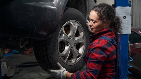 A female mechanic in a red plaid shirt inspects a car tire in a garage.
