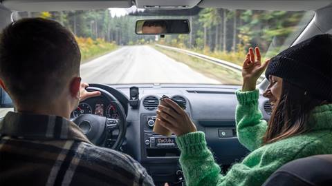 Young woman wearing a green sweater dances in the passenger seat while a man drives; both enjoying a road trip through a forested area.