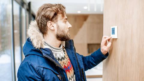 A man in a winter coat adjusts a thermostat on a wooden wall inside a modern building.