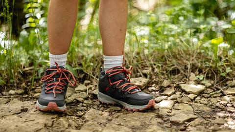 Close-up of a person's legs wearing hiking boots and white socks, standing on a rocky trail surrounded by greenery.
