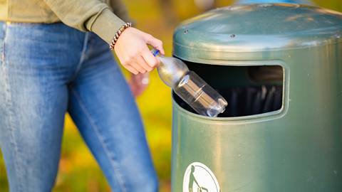 A person recycling a plastic bottle in a designated green bin in a park.