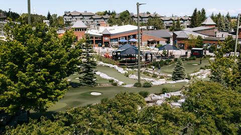 Aerial view of a busy outdoor mini-golf course surrounded by a restaurant and residential buildings on a sunny day.