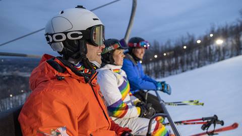 Three skiers wearing helmets and goggles ride a chairlift up a snowy mountain slope during twilight.