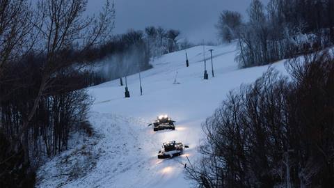 Two snowcats grooming a ski slope at dusk, surrounded by snow-covered trees and mist, with overhead lights illuminating their path.