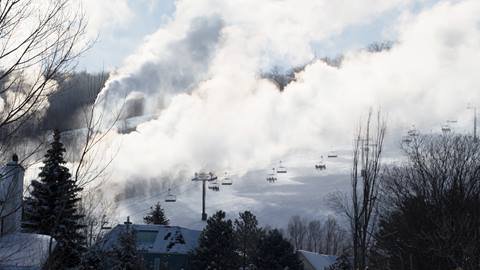 Snow-covered ski slopes with active snow-making machines creating clouds of artificial snow, ski lifts and trees visible.