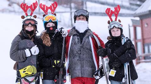 Four people dressed in winter gear and reindeer antlers, one wearing a Santa Claus outfit, stand in the snow with skis and a snowboard.