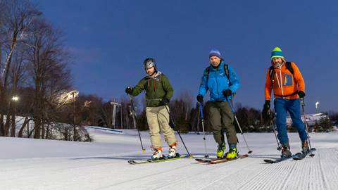 Three people in winter gear and skis walk up a snowy slope under a clear sky.