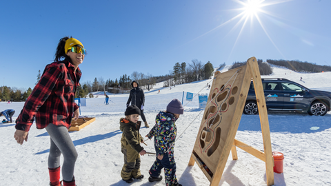 Adults and children play near a snow-covered slope with a wooden board activity under a bright sunlit sky.