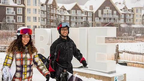 Two people in ski gear walk with snowboards in front of large "BLUE" sign and snow-covered buildings in the background.