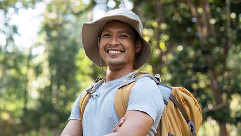 a smiling man with a hat and backpack outdoors in a sunlit forest at blue mountain resort
