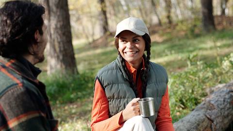 a woman in a cap and vest smiles while holding a metal mug, sitting at a designated picnic area and conversing with a person opposite her at blue mountain resort