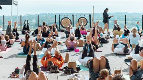 A group of people practice yoga outdoors on mats, raising their legs in the air. A person is standing in front, instructing the class. The backdrop includes a scenic view of the coastline and sea.